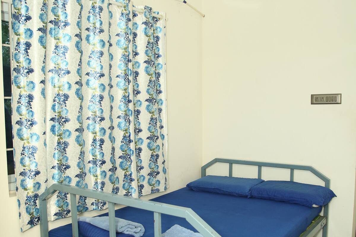 A single bed with a blue mattress and pillows, a wooden chair, a window with floral curtains, and an air conditioning unit.