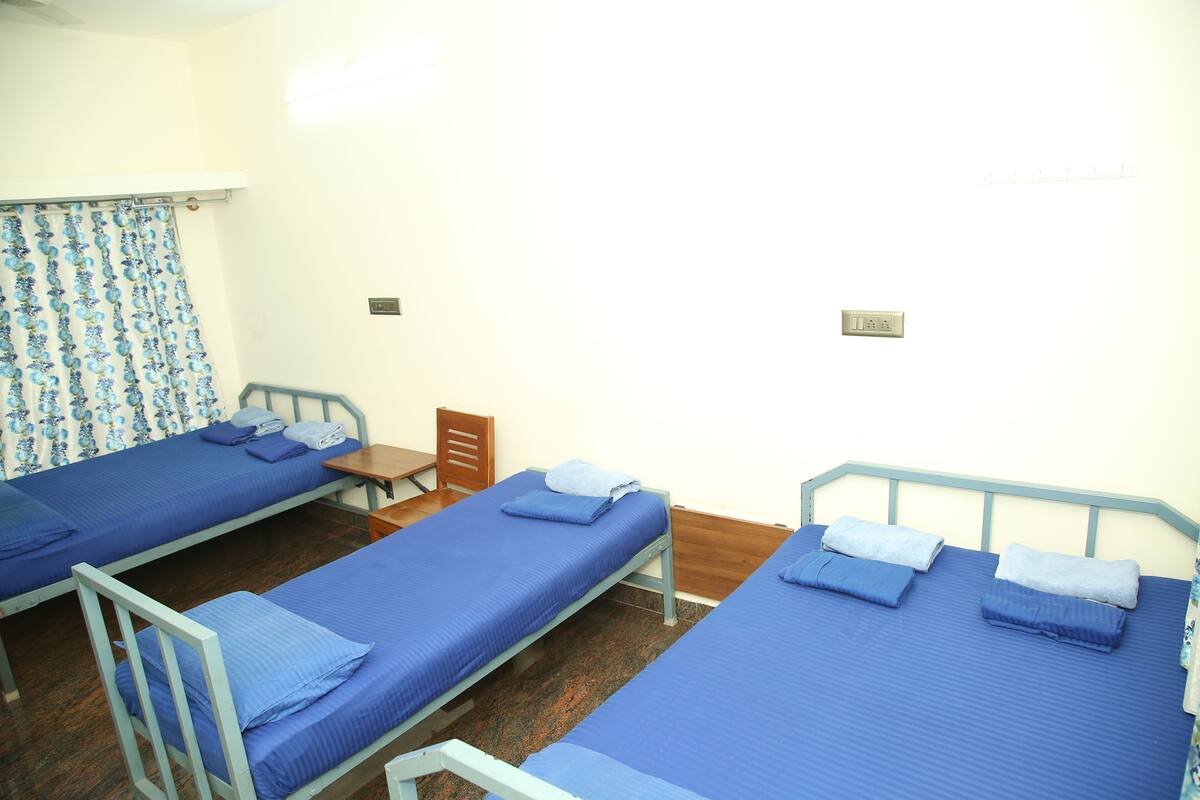 A dormitory room with three single beds, each with blue bedding and pillows. There is a wooden chair and a small table in the room.