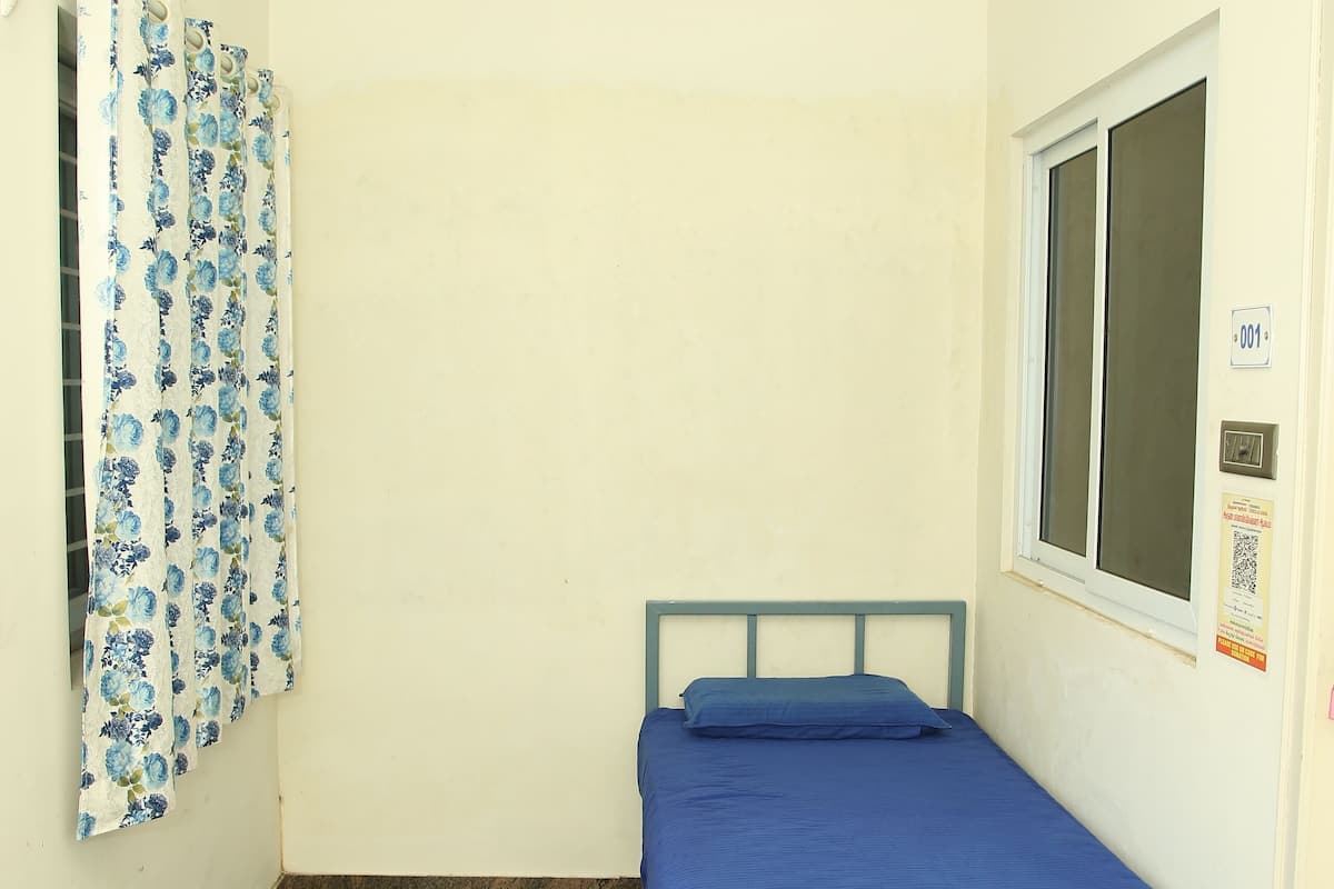 A small, single-bed room with a blue bedspread, a ceiling fan, a window with a view, and a curtain with a floral pattern.