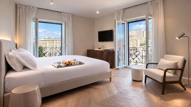 Una camera d'albergo moderna con un letto grande, un angolo salotto con una poltrona, un tavolino e una TV. La stanza dispone di un balcone con vista sulla città.