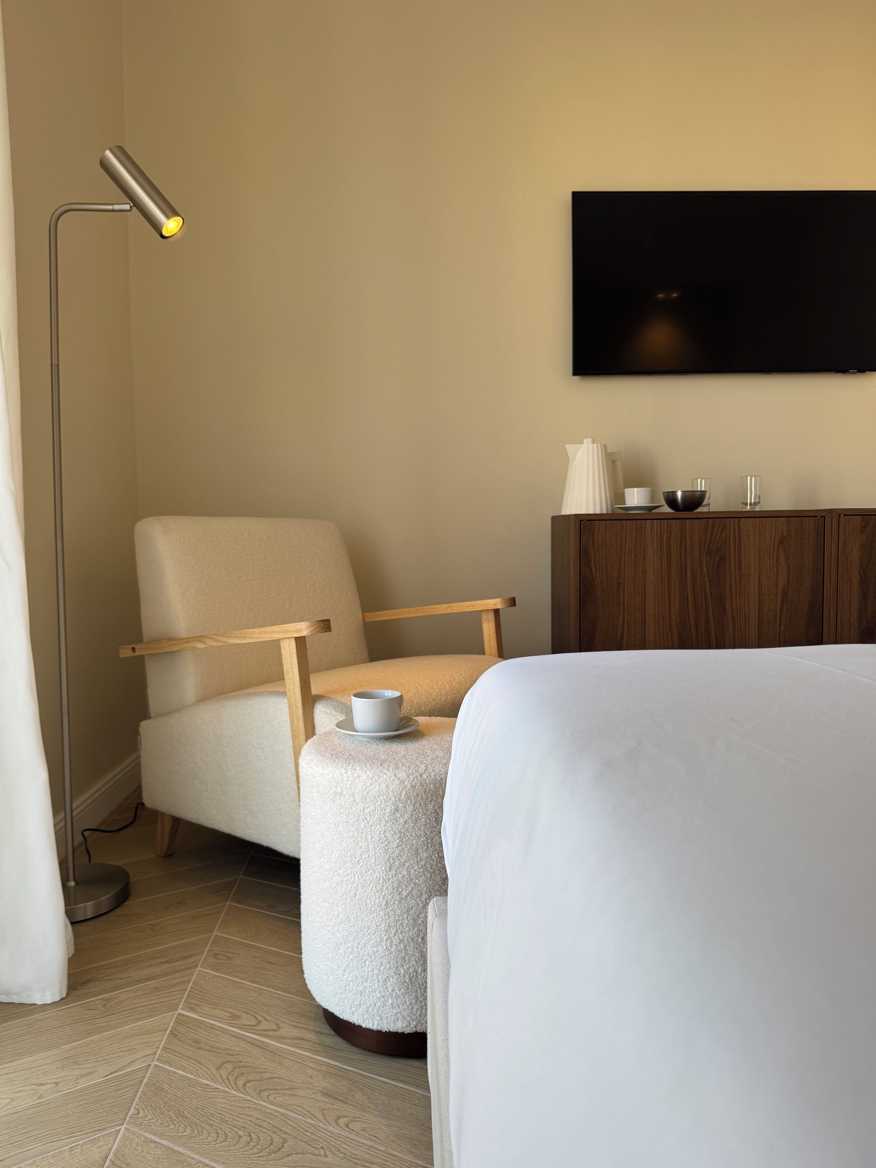 Ein Hotelzimmer mit einem Bett, einem Sessel mit Tasse, einem kleinen Hocker, einem an der Wand befestigten Fernseher und einem Holzschrank.