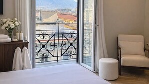 Camera d'hotel con un letto grande, una sedia, un tavolino con articoli da toilette e un balcone con vista sulla città e sulle montagne.