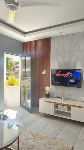 Een moderne woonkamer met een televisie aan een marmeren muur, een wit kastje met goudkleurige handgrepen en een glazen tafel.