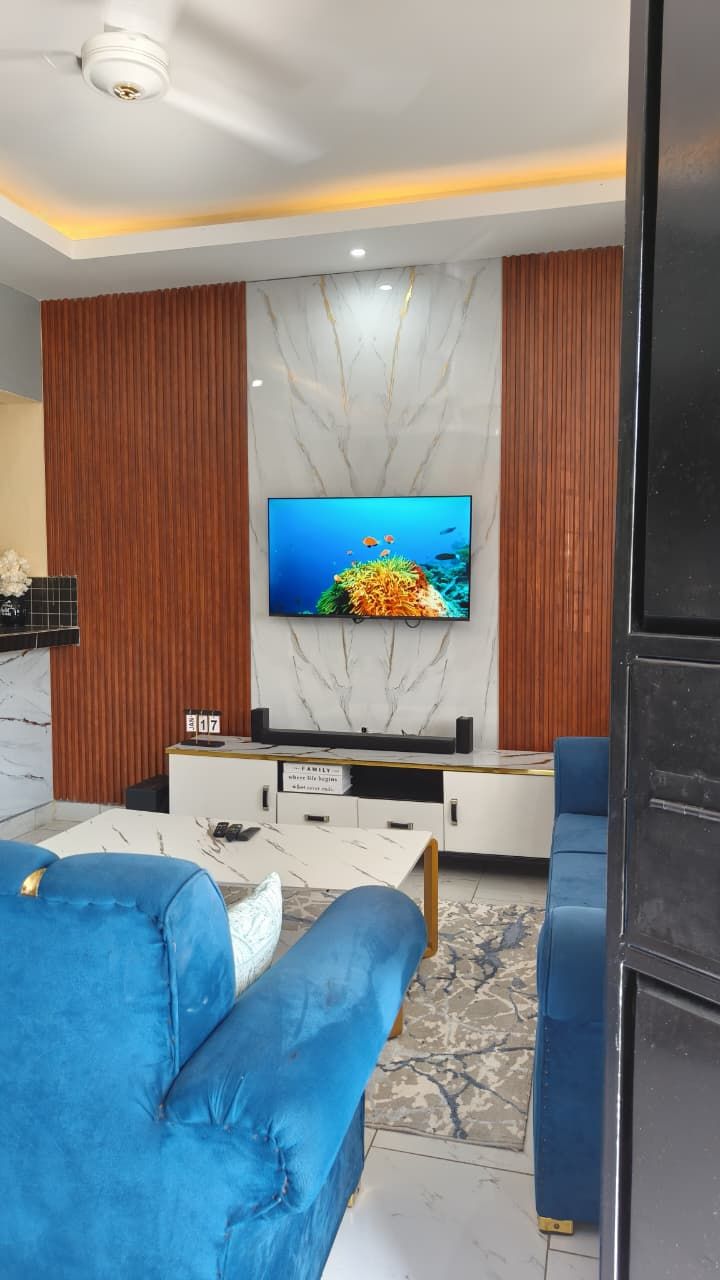A modern living room with a flat-screen TV mounted on a marble wall, blue upholstered sofas, and a wooden accent wall.