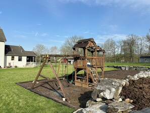 Ein Spielplatz mit einer Holzschaukel in einem grünen Garten, im Hintergrund ein Haus.