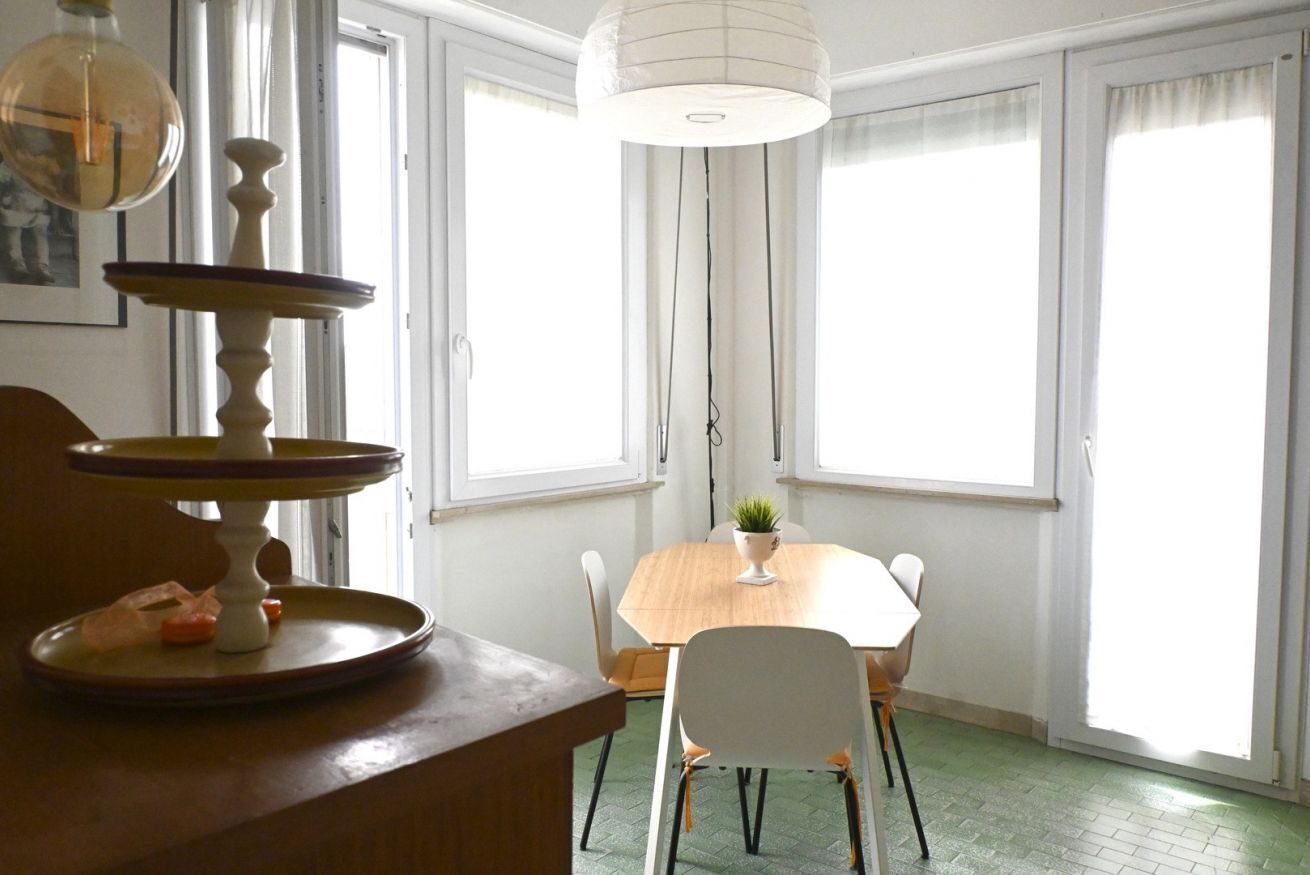 A dining area with a wooden table, white chairs, a hanging light fixture, and a window with curtains.