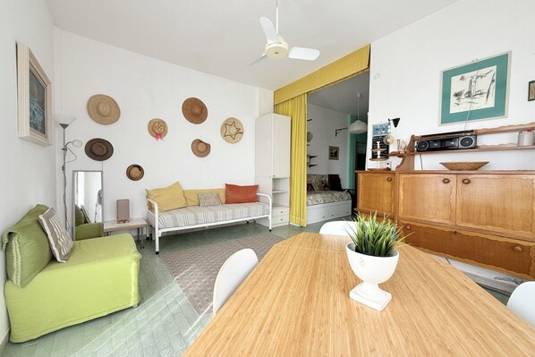 A room with a wooden dining table, a green sofa, a white armchair, a ceiling fan, and a wall with decorative hats.