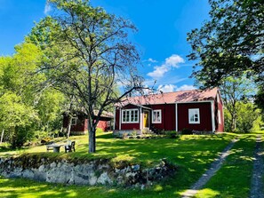 A red wooden house with a stone wall, a bench, and a tree in a sunny garden. - 6 Person Holiday Home in Backaryd (Backaryd)