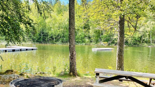 A lakeside picnic area with a concrete grill, a wooden bench, and a dock extending into the water. - 6 Person Holiday Home in Backaryd (Backaryd)