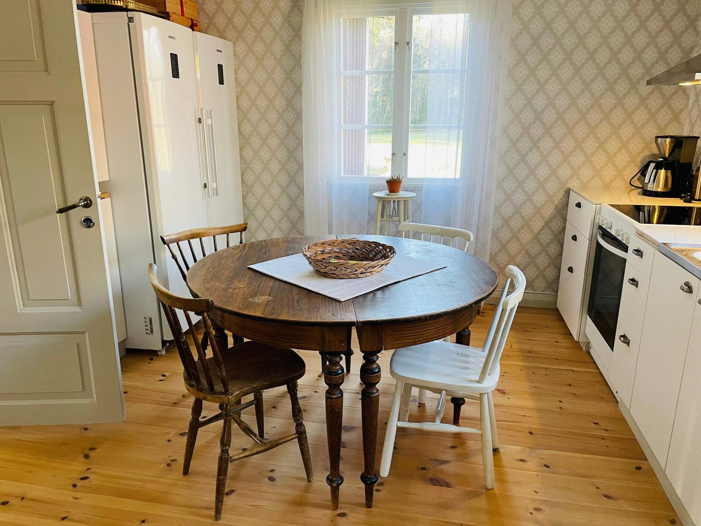 A dining area with a round wooden table, two chairs, a white cabinet, and a window with curtains.