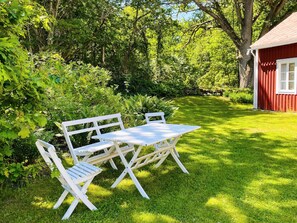 A white wooden table and chairs set on a lush green lawn with dense foliage and a red building in the background. - 6 Person Holiday Home in Backaryd (Backaryd)