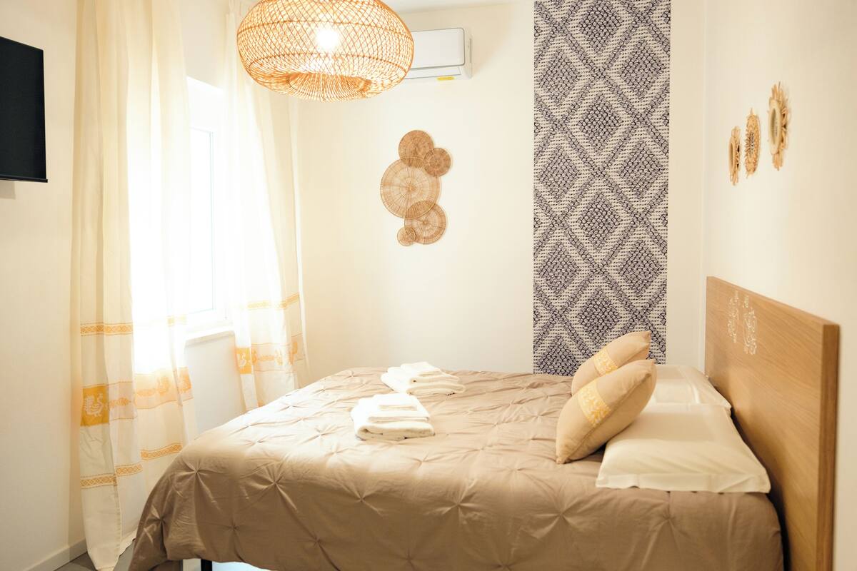 A neatly made bed with a beige comforter and pillows, a wall-mounted air conditioner, a patterned wall, and a woven light fixture.