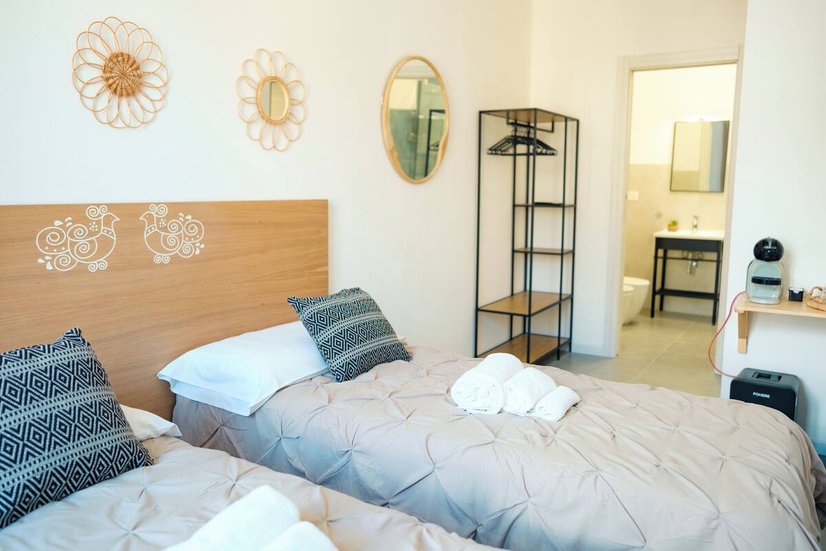 A hotel room with two beds, a wooden headboard, patterned pillows, a mirror, a shelf with a coffee maker, and a folded towel on the bed.