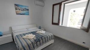 Quarto com cama, mesa de cabeceira, abajur, pintura e janela.
