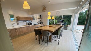 Une cuisine moderne avec des meubles en bois, une grande table à manger, des luminaires suspendus et une vue sur le jardin grâce à de grandes fenêtres.