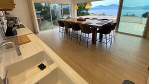 Une cuisine moderne dotée d’un grand îlot central, d’un coin repas avec des chaises, d’un évier et offrant une vue sur la mer.