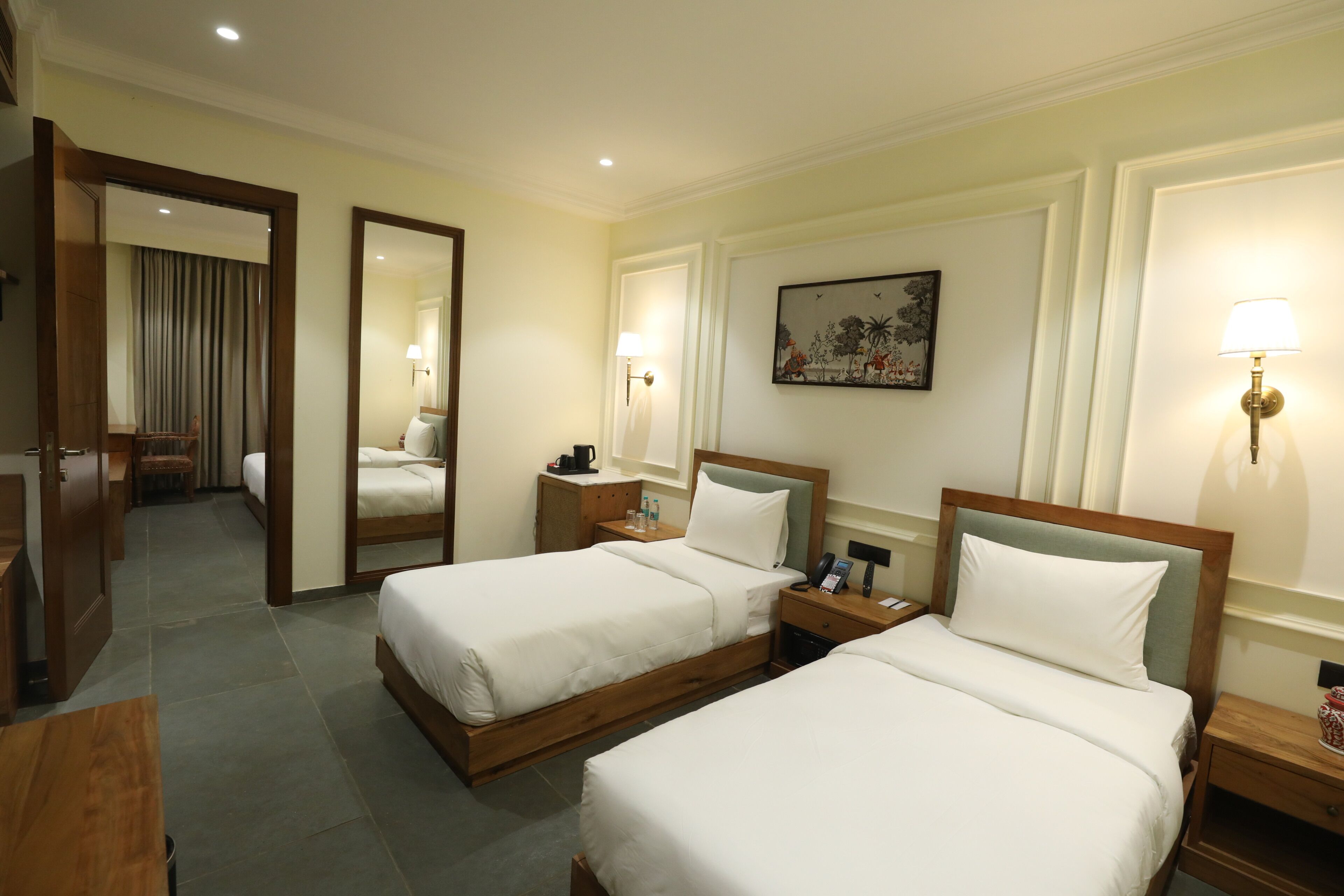 A hotel room with two beds, a desk, a chair, a mirror, a nightstand, a lamp, a painting, and a door leading to another room.