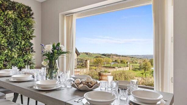 Una mesa de comedor dispuesta con platos, vasos y un bol de frutos secos, con vistas a un paisaje pintoresco.