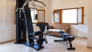 A home gym with a weight bench, a squat rack, and a bench press. There is a wicker sofa and a window with wooden frames. - Studio for Couples in Capo Vaticano With Pool (Ricadi)
