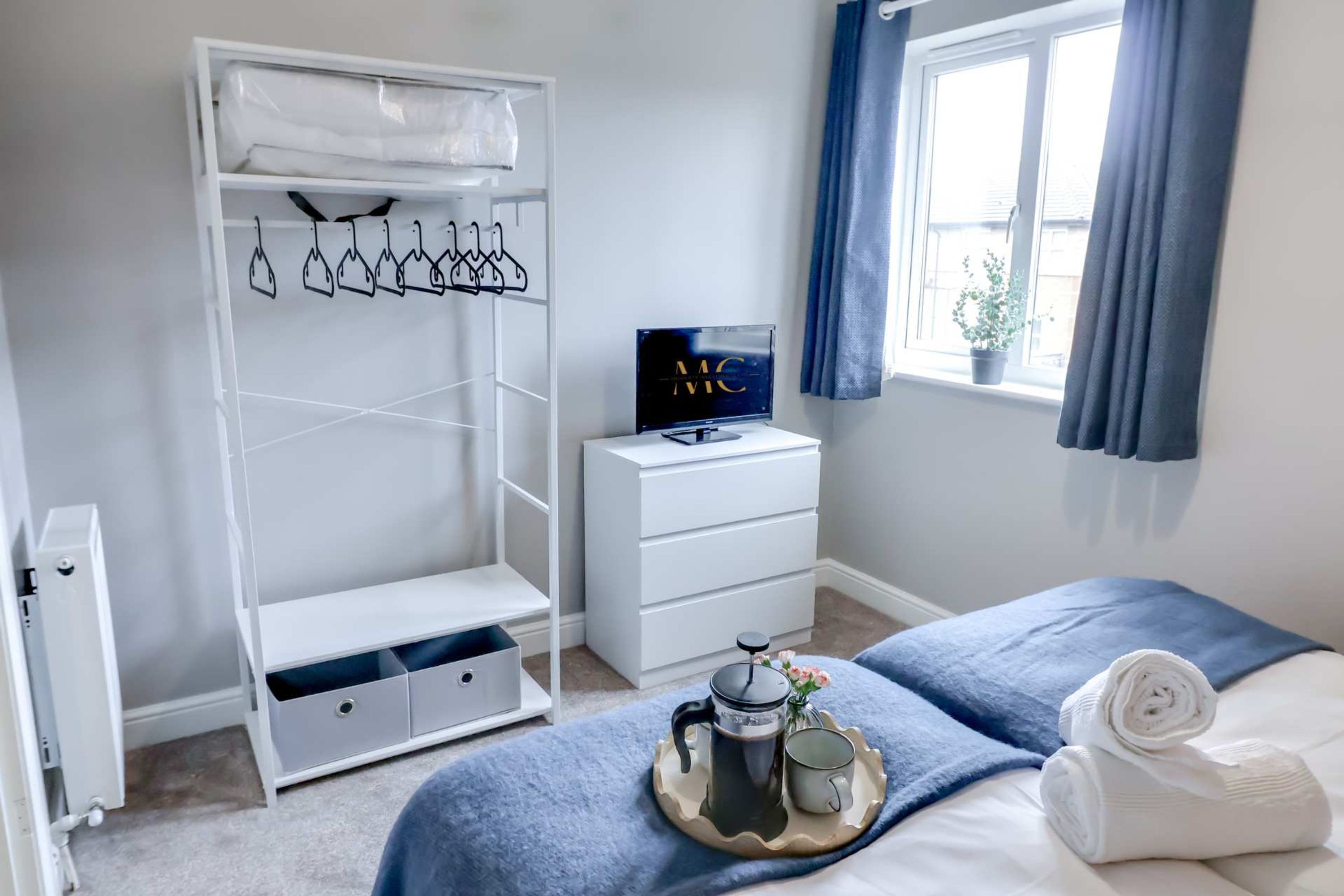 A neatly organized bedroom with a bed, a bedside table with a coffee maker and mug, a white wardrobe, a TV, and a window with curtains.