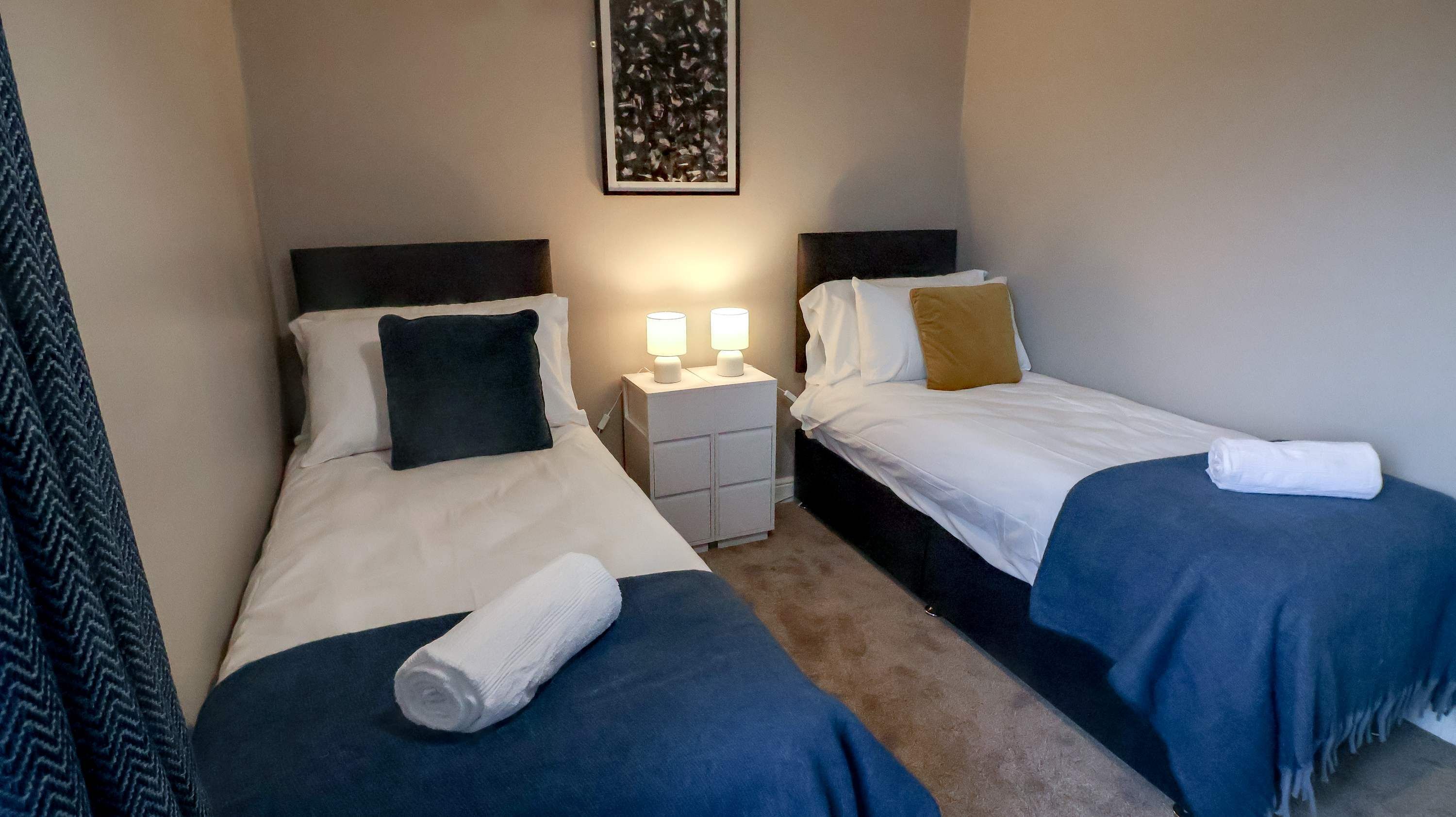 Ein Hotelzimmer mit zwei Betten, jeweils mit einem Kopfkissen und einem gerollten Handtuch. Dazwischen befindet sich ein Nachttisch mit zwei Lampen.