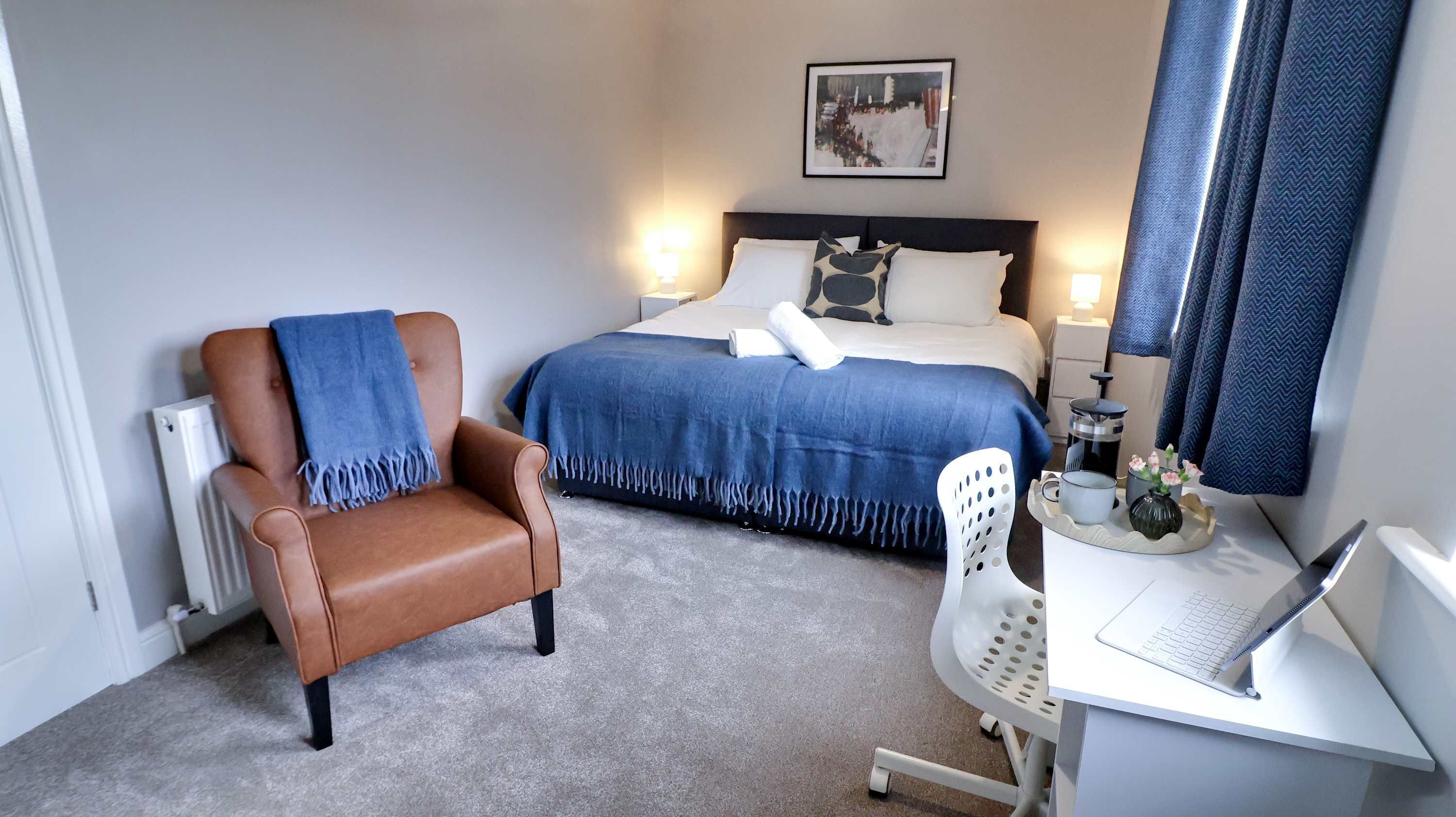 A hotel room with a bed, a chair, a desk with a laptop, a radiator, and a wall-mounted picture.