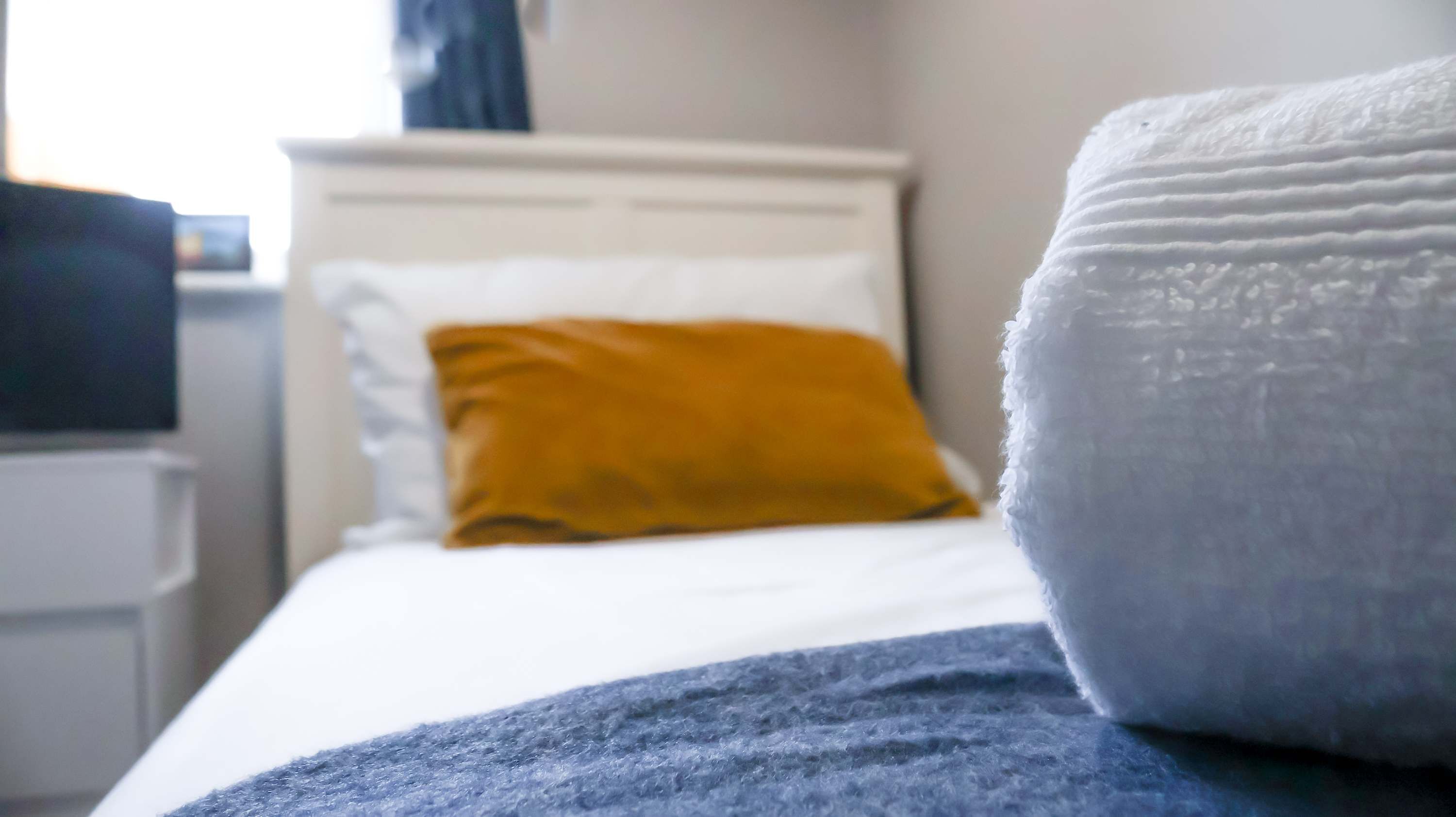 Een bed met witte en mosterdkleurige kussens, een blauwe deken en een witte hoofdbord.