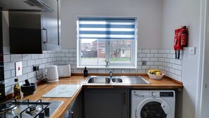 A compact kitchen with a built-in washing machine, a coffee maker, and a window with blinds. - Modern House In Hartlepool With Private Parking (Hartlepool)