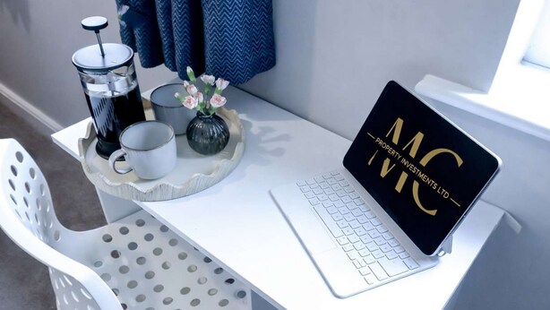 A white desk with a laptop displaying "MC Property Investments Ltd" logo, a coffee maker, a mug, and a vase with flowers. - Modern House In Hartlepool With Private Parking (Hartlepool)