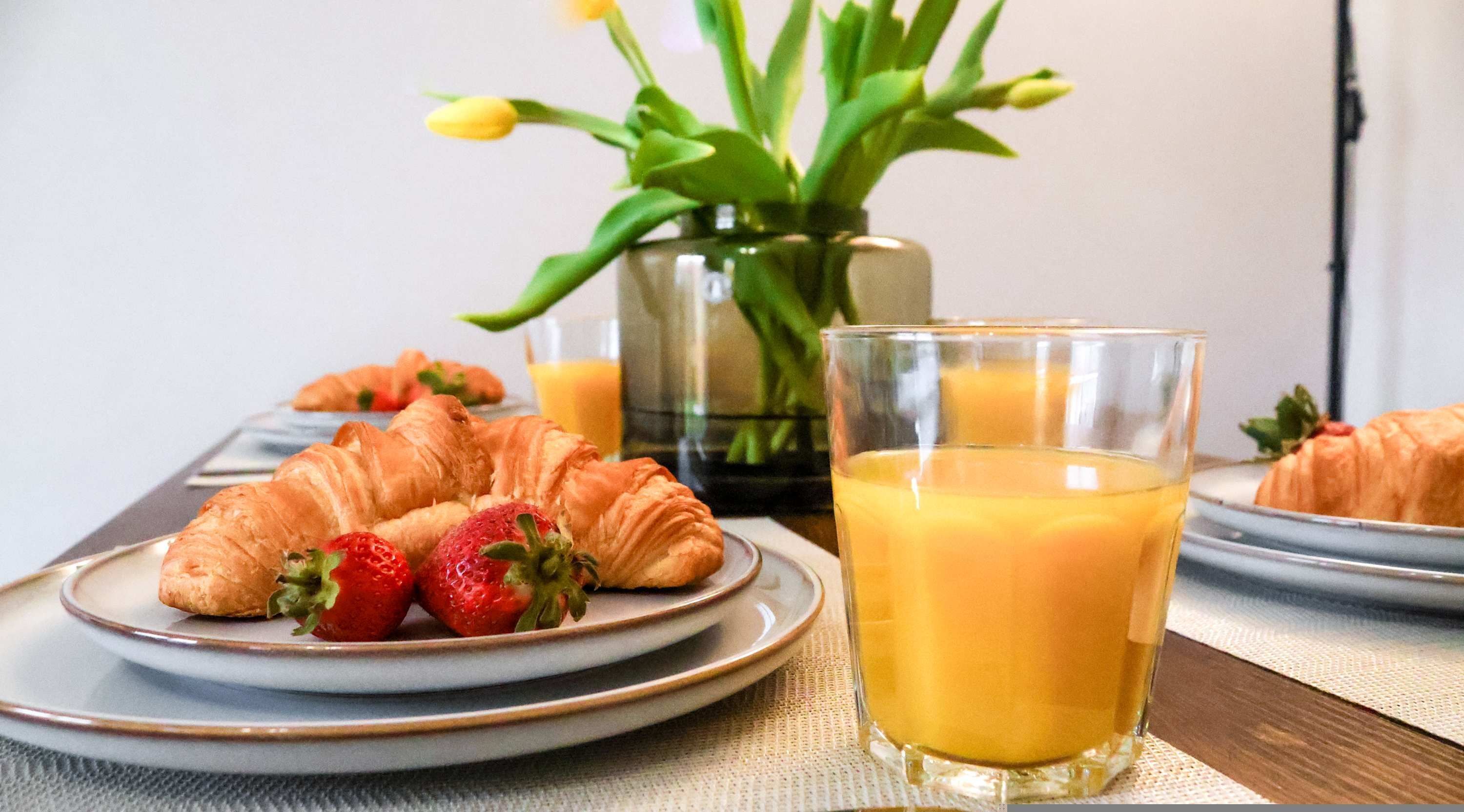 Un desayuno con cruasanes, fresas y zumo de naranja en platos, con un jarrón de flores al fondo.
