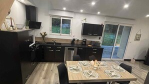 Une cuisine moderne avec des meubles de couleur sombre, un coin repas avec une table en bois dressée pour quatre personnes, et une grande fenêtre offrant une vue sur l’extérieur.