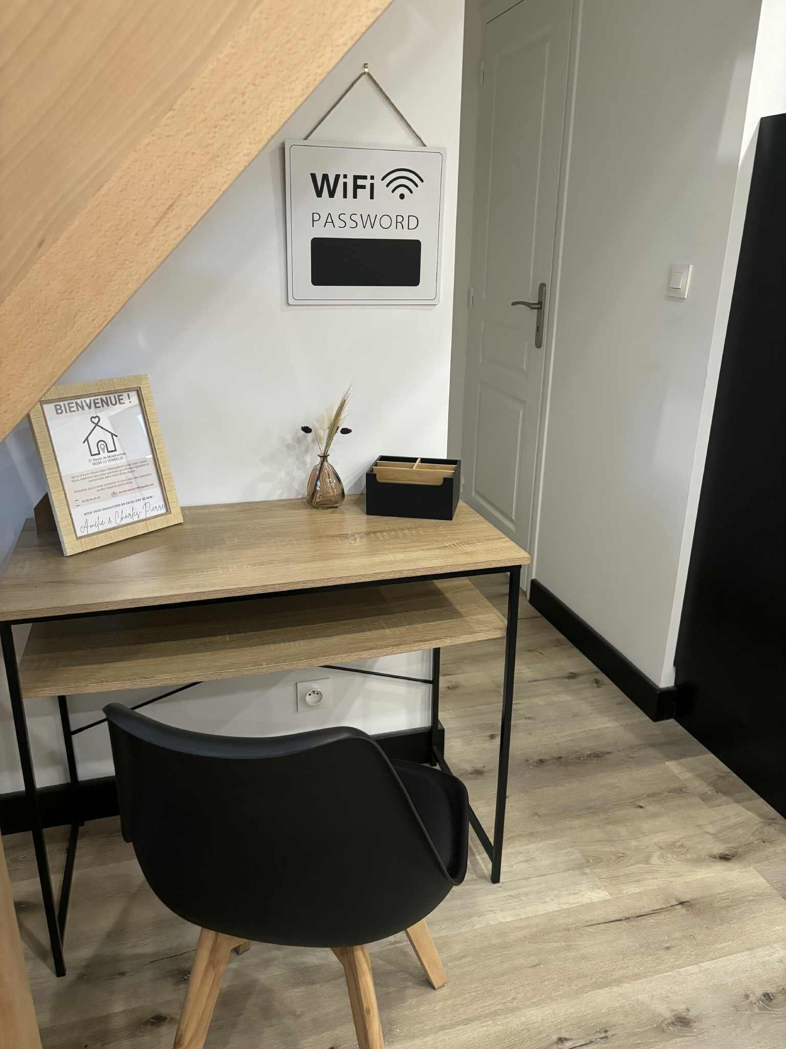 A modern workspace with a wooden desk, a black chair, a framed welcome sign, a vase with dried plants, and a WiFi password sign on the wall.