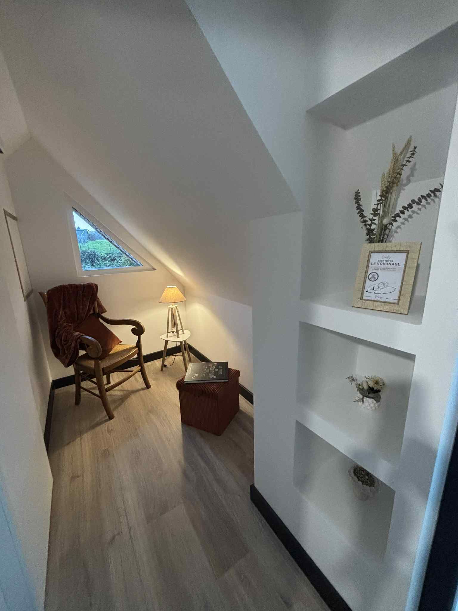 Una camera accogliente con soffitto a falde, una poltrona a dondolo in legno, un tavolino e una lampada. Si vede del verde attraverso la finestra.