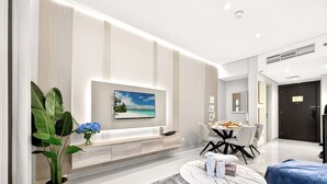 A modern living room with a flat-screen TV mounted on the wall, a dining area with a round table and chairs, and a large potted plant. - WYZ RENT|Grand 1BR Cour Jardin Near Bay Avenue (Dubai)
