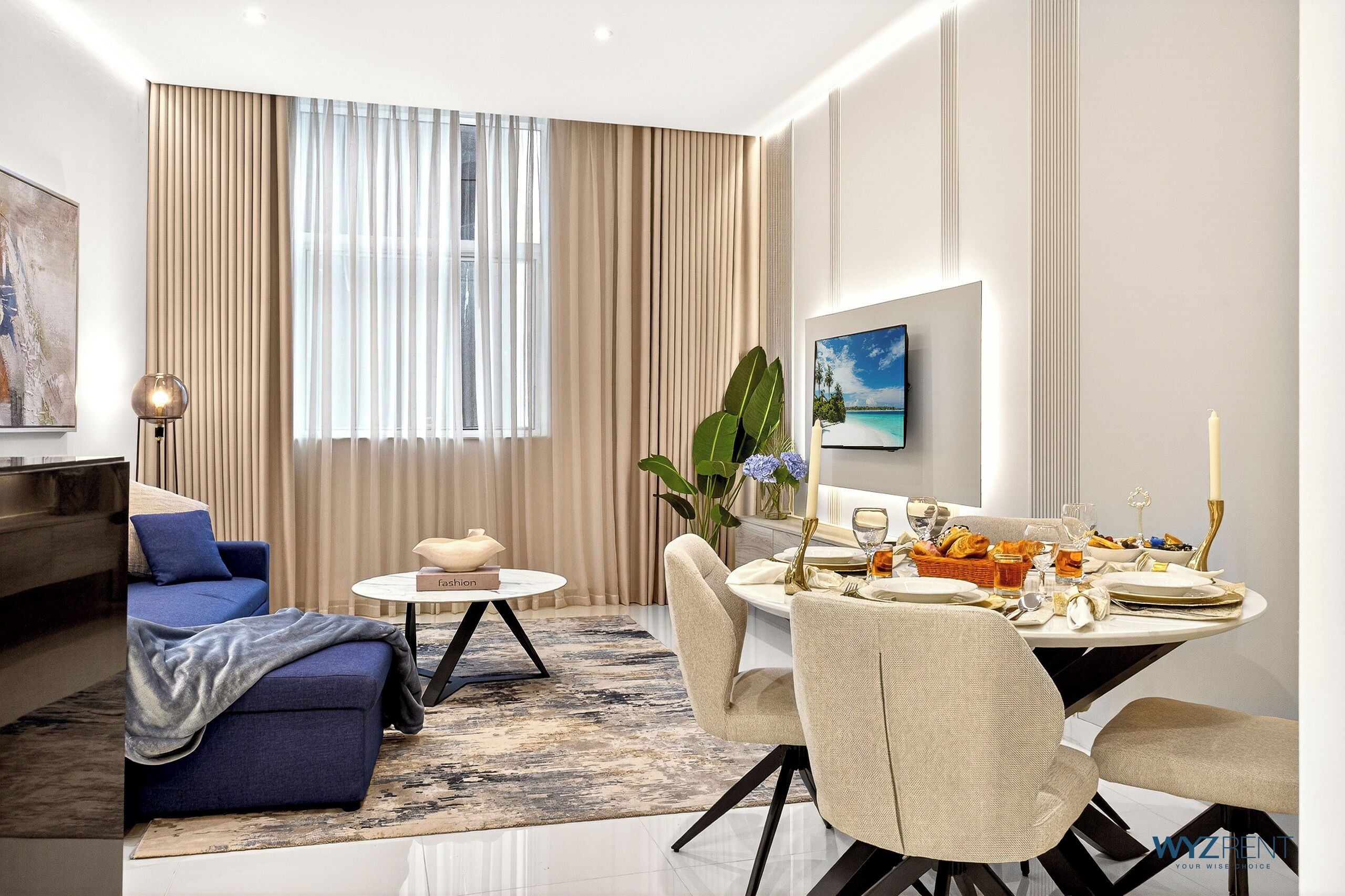 Una sala de estar moderna con un sofá azul, una mesa de comedor redonda para cuatro personas y un amplio ventanal con cortinas color beige.