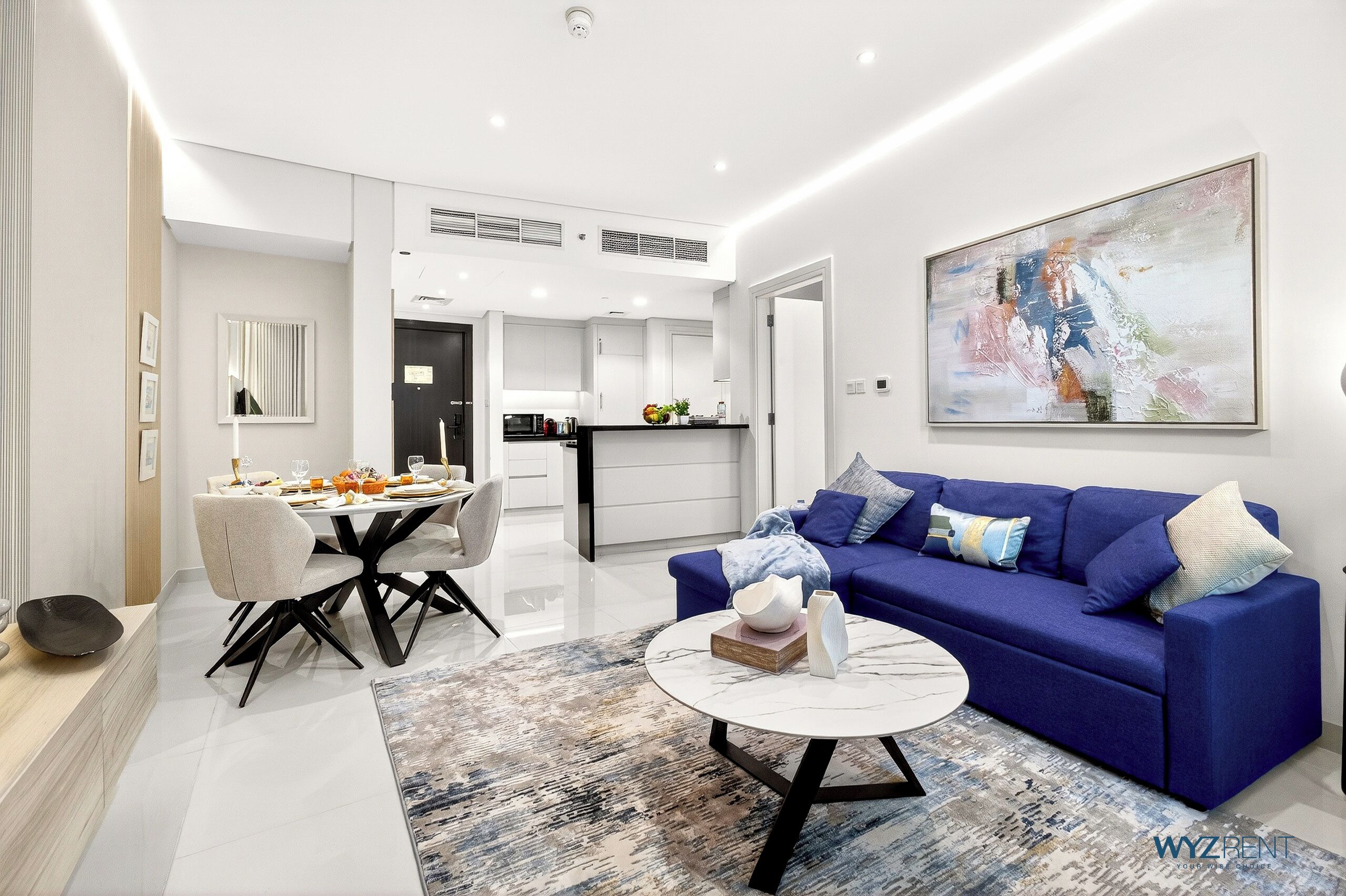 A modern living room with a blue sofa, a white coffee table, and a dining area with a white table and chairs.