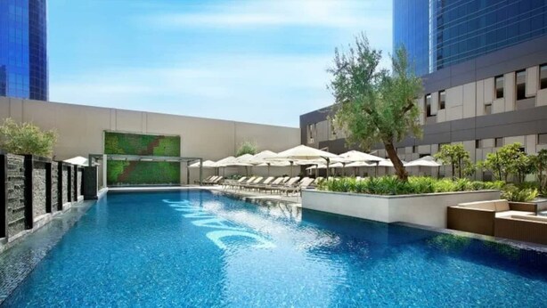 A swimming pool with lounge chairs and umbrellas, surrounded by modern buildings. - WYZ RENT|Grand 1BR Cour Jardin Near Bay Avenue (Dubai)