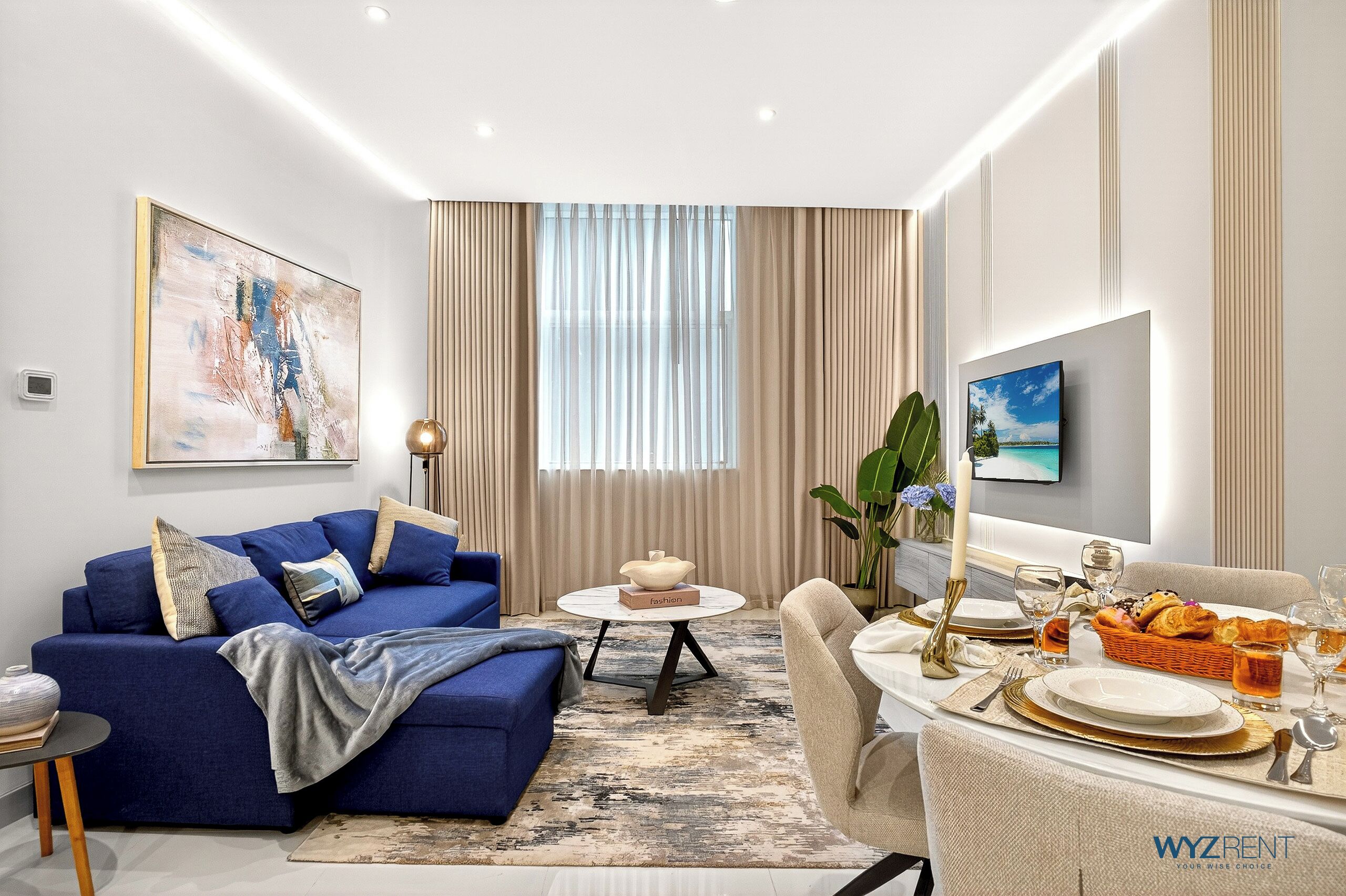 A modern living room with a blue sofa, a dining table set for a meal, and a large window with curtains.