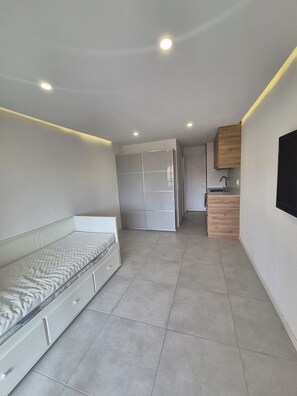 Una habitación moderna y compacta con una cama, un armario, una pequeña kitchenette y un televisor montado en la pared.