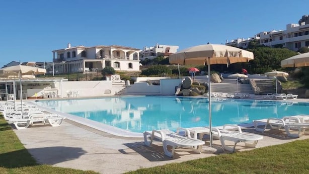 En swimmingpool med solstolar, parasoller och byggnader i bakgrunden.