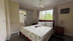 Een slaapkamer met een bed, een plafondventilator, een raam met gordijnen, een nachtkastje en een deur naar een badkamer.