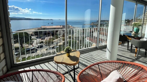 Ein Balkon mit Blick auf das Meer und die Stadtlandschaft, mit Korbmöbeln, einem kleinen Tisch und einem Blumentopf.
