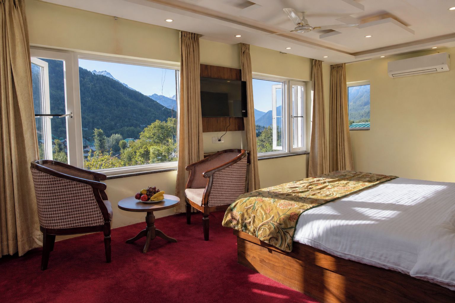 Habitación de hotel con cama, dos sillas, una mesita con fruta y vistas a montañas a través de amplios ventanales.
