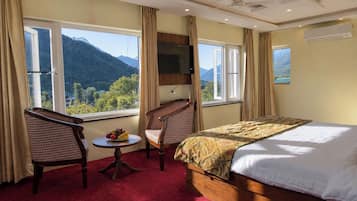 A hotel room with a bed, two chairs, a small table with fruit, and a view of mountains through large windows.