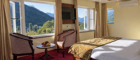 A hotel room with a bed, two chairs, a small table with fruit, and a view of mountains through large windows.