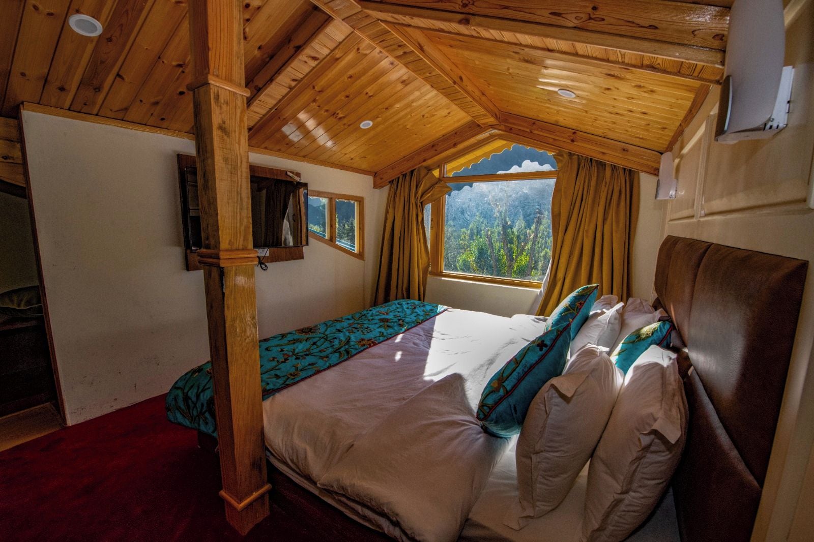 A bedroom with a wooden ceiling, a bed with multiple pillows, a window with curtains, and a view of trees.