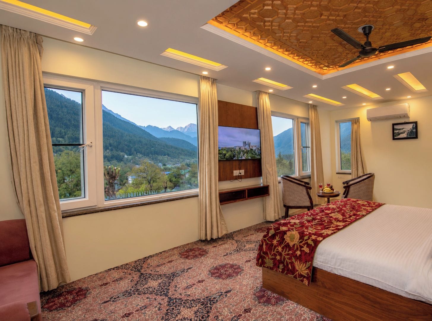 A hotel room with a bed, a TV, a desk, and a chair. There are large windows with curtains, and a mountain view is visible outside.