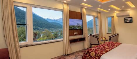 Habitación de hotel con cama, televisión, escritorio y silla. Amplias ventanas con cortinas y vistas a la montaña.
