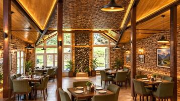 A restaurant with high ceilings, wooden beams, and a stone wall. Tables are set with chairs, and there are plants and artwork on the walls.