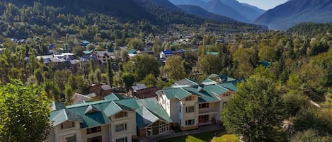 Un edificio de varios pisos con techo verde, rodeado de abundante vegetación y montañas al fondo.